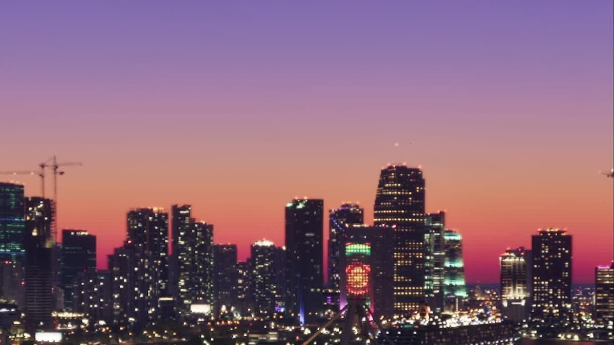 Skyline de Vice City ao entardecer, com prédios iluminados e guindastes de obra ao fundo